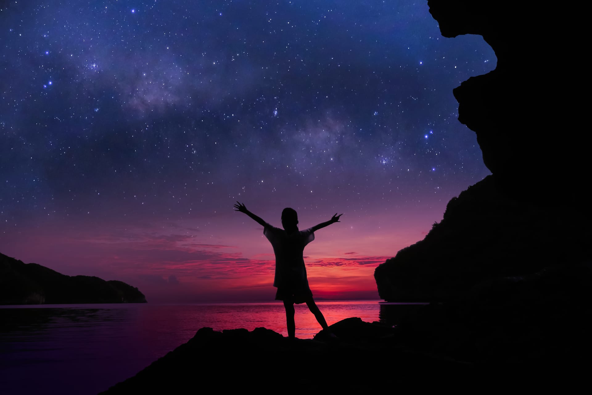 Girl at night on top of rock near ocean looking at starry sky with arms outstretched