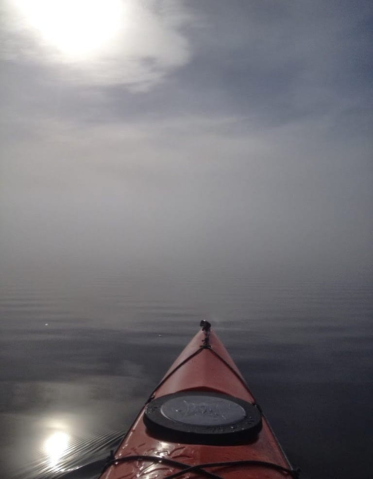 Photo of front tip of orange kayak on calm lagoon with the sun and bright blur in the clouds and reflected on the water.