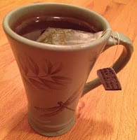 Photo of tall green cup with a dragonfly on it filled with tea, the teabag tag hanging over the lip of the cup.