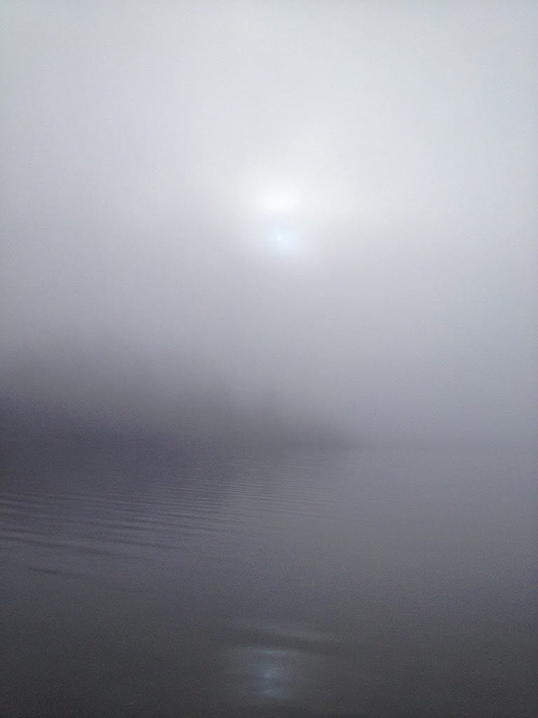 Photo of misty, foggy calm lagoon and grey sky, the foggy outline of trees in the distance and the heavily muted sun in the clouds and reflected on the water.