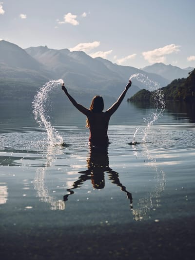 Photo of woman waist-deep in river flinging water joyfulling into air, reflected in water.