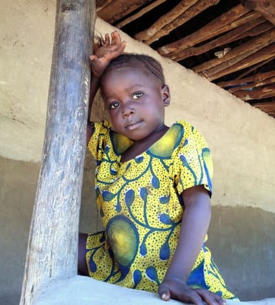 Photo of a little girl in rural Sierra Leone wearing bright yellow and blue country-cloth dress, standing outside her painted clay home.