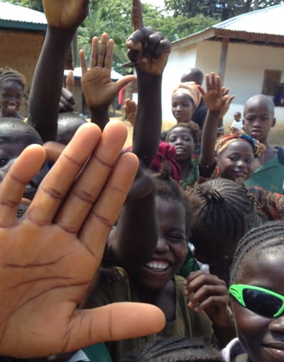 Photo of a group of children in a rural Sierra Leone village, smiling, hands raised for a 'high-5.'